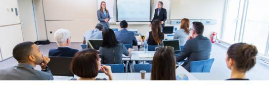 一群穿着商务休闲装的员工坐在教室里，背对着镜头，面对着站在教室前面、投影仪屏幕两侧的两位老师